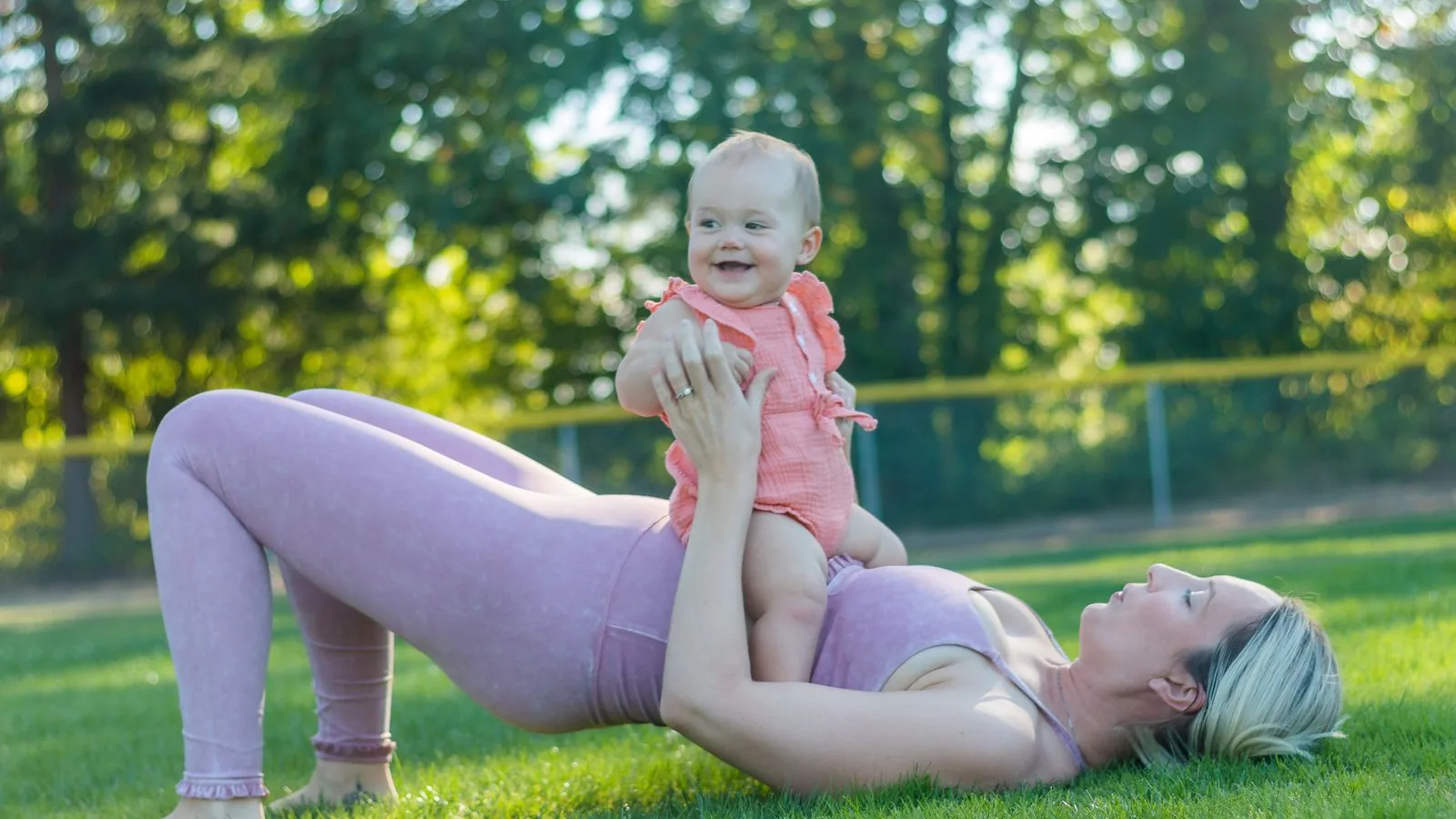 Mutter liegt auf einer Wiese in der Brückenposition und hält ihr lachendes Baby über sich