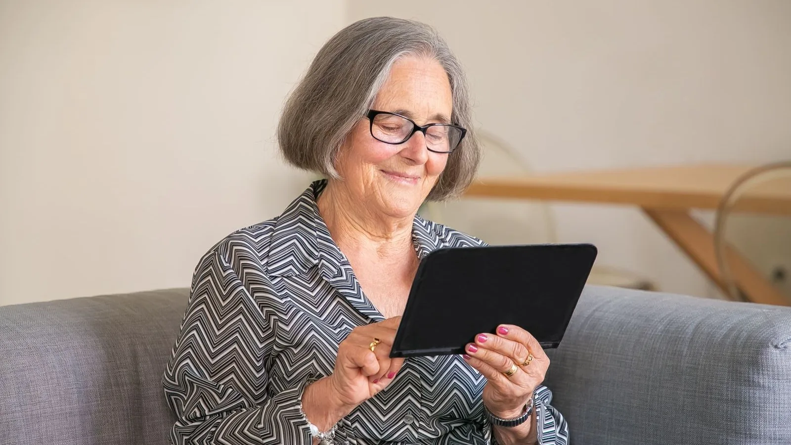 Ältere Frau liest lächelnd auf einem Tablet auf dem Sofa