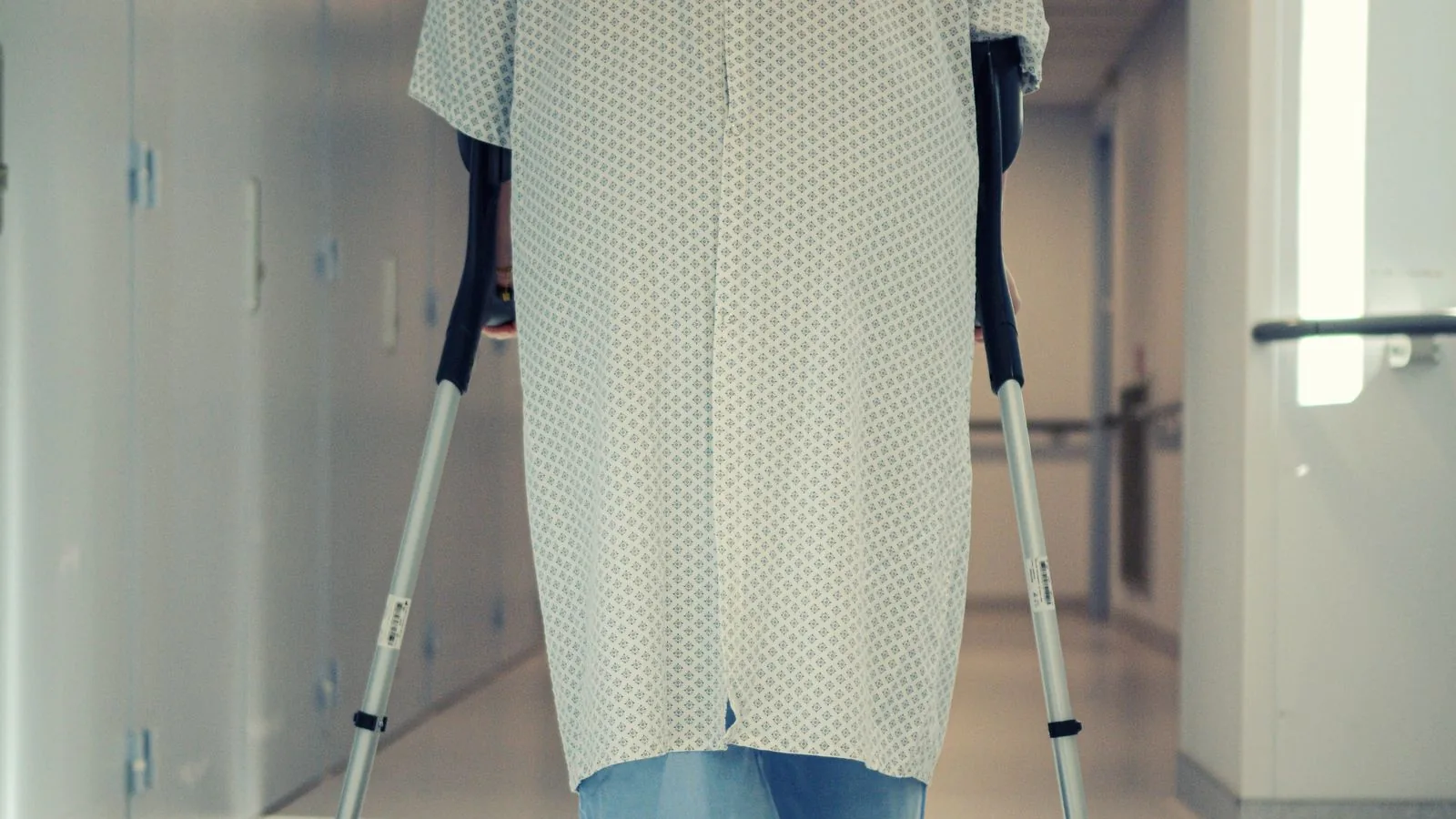 Patient geht mit Unterarmgehstützen durch einen Klinikflur, Rückenansicht